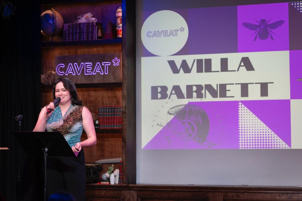 Willa on stage at Caveat NYC with a microphone, a projected slide reading Caveat and Willa Barnett behind her